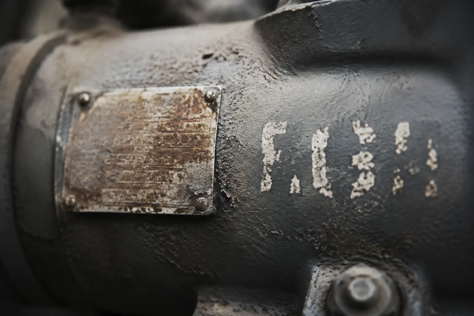a close view of a pump casing carrying two different identity marks side by side, a corroded riveted metal manufacturer nameplate and a hand-stencilled equipment number painted directly onto the casing in a different convention