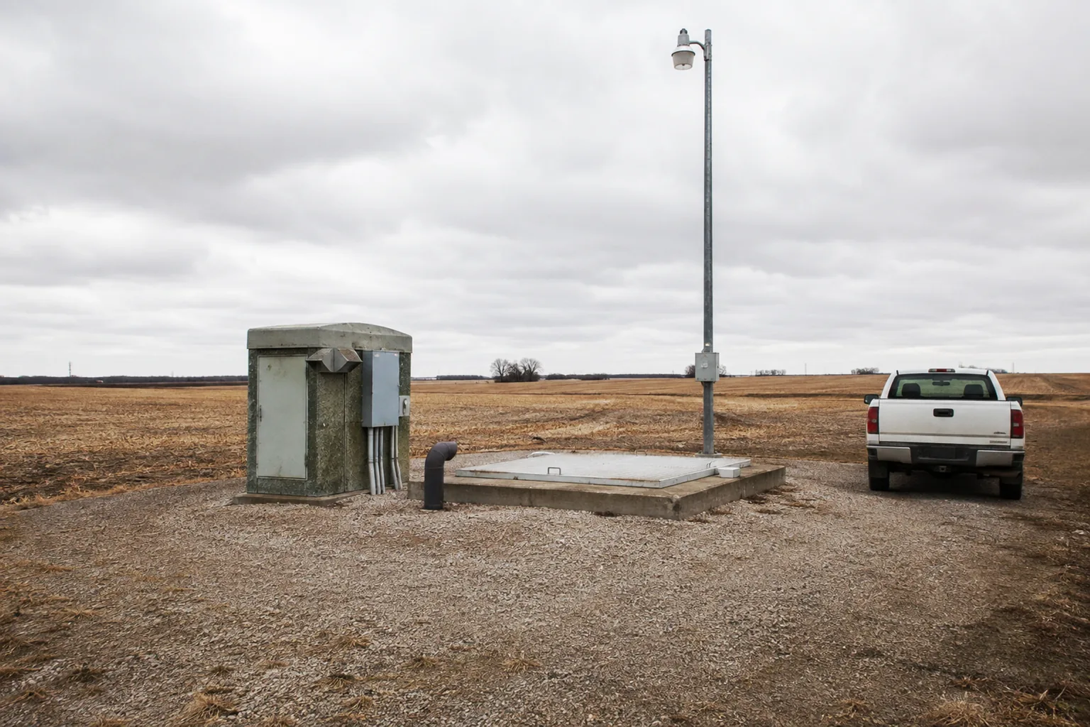 a small rural sewage lift station on a gravel apron at the edge of a field, a modest control kiosk, a concrete hatch, a vent stack and one pole light, a single pickup truck parked alongside under an overcast sky