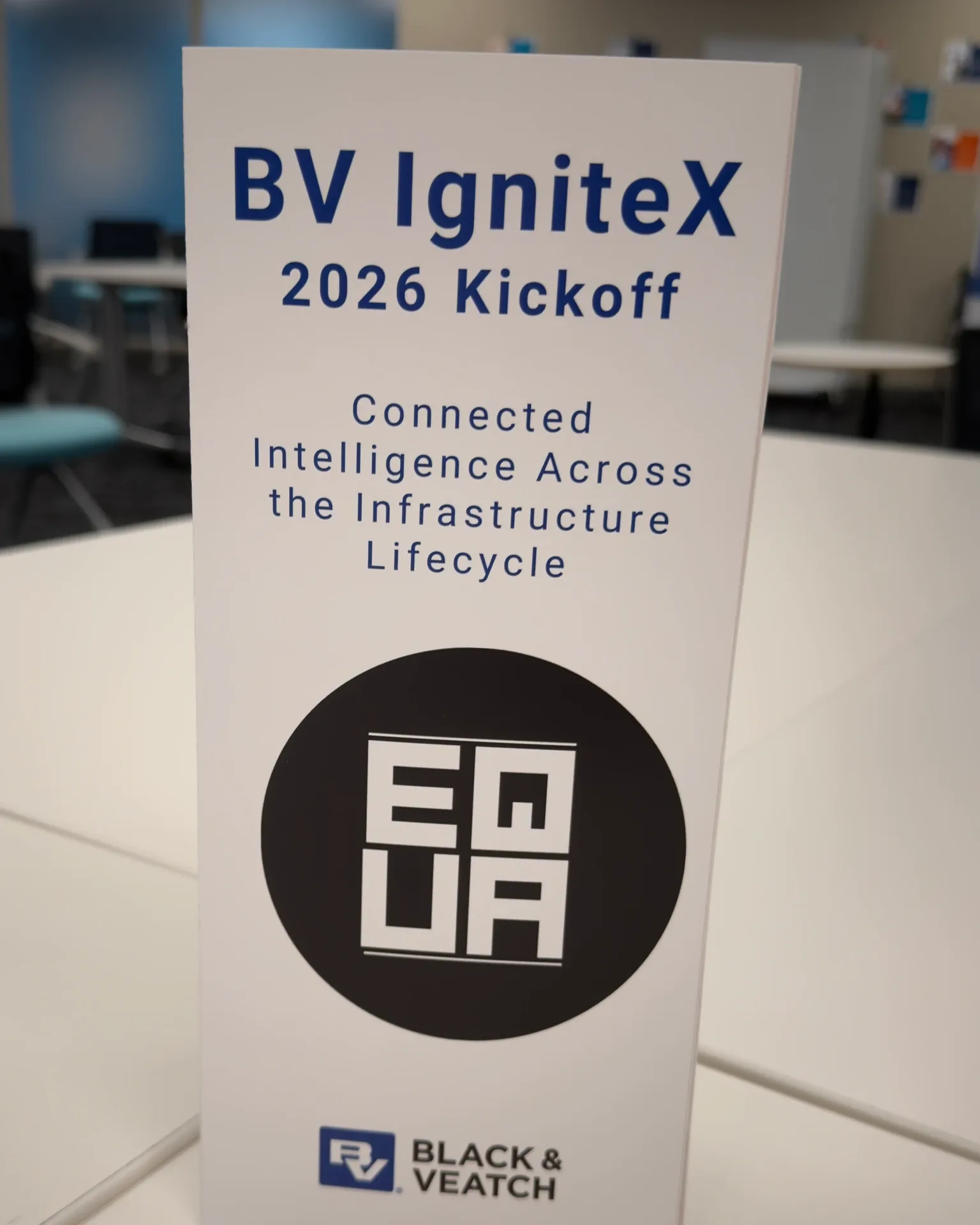 A printed tabletop sign reading BV IgniteX 2026 Kickoff, Connected Intelligence Across the Infrastructure Lifecycle, carrying the EQUA circular mark above the Black and Veatch logo, on a white table in a meeting room